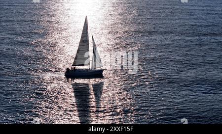 Ein kleines Segelboot gleitet anmutig über glitzerndes Wasser und reflektiert die warmen Sonnenuntergänge, während die Crew einen ruhigen Moment auf dem offenen Meer genießt. Stockfoto