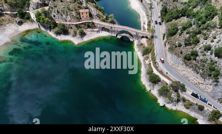 Das ruhige Wasser eines Sees glitzert unter der Sonne, mit einer rustikalen Brücke, die felsige Ufer verbindet, und einem malerischen Gebäude inmitten von Grün. Stockfoto