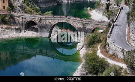 Unter einem hellen Himmel überspannt eine zeitlose Steinbrücke einen glitzernden grünen Fluss, während Besucher entlang der Ufer schlendern und die ruhige Schönheit von Th genießen Stockfoto