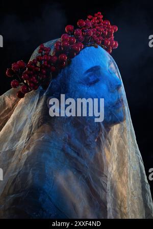 Ein Mädchen mit wunderschönem Make-up, ihr Gesicht ist bedeckt mit blauem Tüll, ein Kranz voller Beeren auf dem Kopf. Halloween-Konzept. Stockfoto