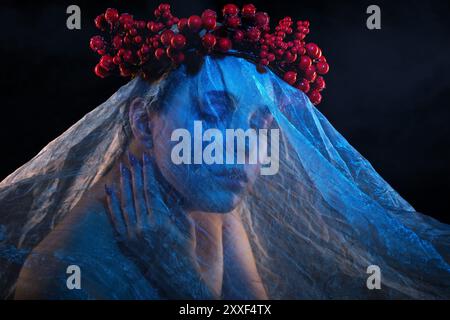 Ein Mädchen mit wunderschönem Make-up, ihr Gesicht ist bedeckt mit blauem Tüll, ein Kranz voller Beeren auf dem Kopf. Halloween-Konzept. Stockfoto