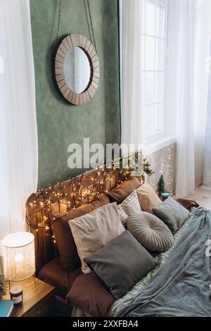 Festlich eingerichtetes Bett mit Holzkopfteil, Feenlichtern und Kissen, das durch große Fenster mit weißen Vorhängen an der grünen Wand positioniert wird Stockfoto