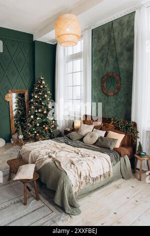 Elegantes Schlafzimmer mit weihnachtlicher Einrichtung, großem Baum, gemütlichem Bett mit Kissen und Decken, Holzkommode, Ganzkörperspiegel und großen Fenstern Stockfoto