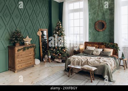 Festliches Schlafzimmer mit Weihnachtsbaum, gemütlichem Bett, Holzkommode, Spiegel in voller Länge und großen Fenstern. Grüne Wände und Weihnachtsdekorationen sorgen für Wärme Stockfoto