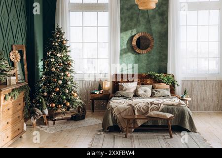 Helles Schlafzimmer mit großen Fenstern, Weihnachtsbaum, gemütlichem Bett, Holzmöbeln und festlicher Dekoration. Grüne Wände und Sonnenlicht sorgen für eine angenehme Atmosphäre Stockfoto