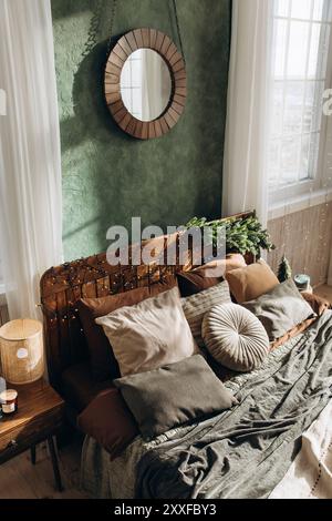 Holzbett mit Lichterketten und Kissen in gemütlichem Schlafzimmer. Sonnenlicht durch Fenster beleuchtet die grüne Wand und den runden Spiegel über dem Kopfteil Stockfoto