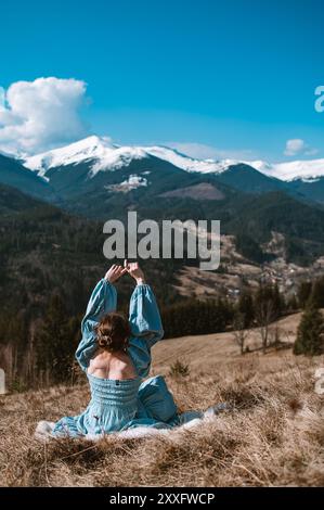 Frau in blauem Kleid genießt während der Frühlings- oder Herbstsaison den schneebedeckten Blick auf die Berge und sitzt auf dem gelben Gras. Gefangen in den Karpaten Stockfoto