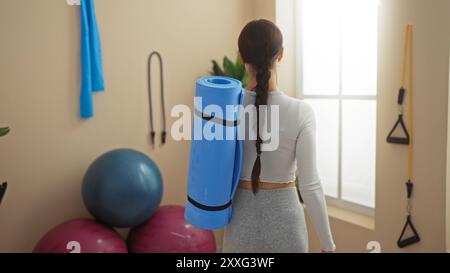 Frau mit blauer Yogamatte im Fitnessstudio mit Fitnessgeräten, weißem Oberteil und grauer Leggings Stockfoto