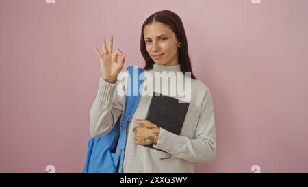 Junge hispanische Frau, die ein Notizbuch hält, das eine gute Geste über isoliertem rosa Hintergrund zeigt Stockfoto