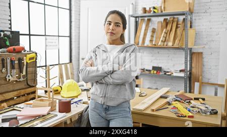 Selbstbewusste junge Frau mit überkreuzten Armen steht in einer Tischlerei, umgeben von Werkzeugen und Holz. Stockfoto