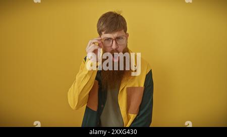Ein überraschter bärtiger junger Mann mit Brille und einem bunten Hemd auf einem isolierten gelben Hintergrund. Stockfoto