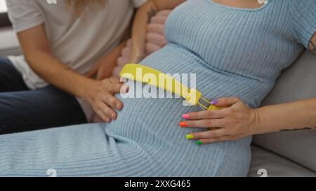 Schwangere Frau mit bunten Nägeln und gelbem Gürtel, die Musik auf ihrem Bauch spielt, begleitet von Mann mit Bart in einem gemütlichen Wohnzimmer, symbolisiert Liebe A Stockfoto