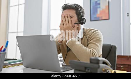 Ein gestresster hispanischer Mann mit Bart und Headset bedeckt sein Gesicht in einem modernen Büro. Stockfoto