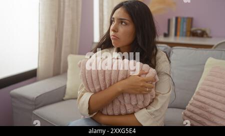 Junge Frau mit langen brünetten Haaren, die in einem Wohnzimmer mit einem rosa Kissen sitzt und ein nachdenkliches und attraktives Verhalten in einer gemütlichen Wohnung ausstrahlt Stockfoto