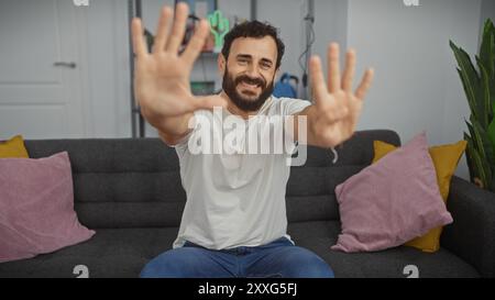 Lächelnder bärtiger Mann in lässiger Kleidung, der die Hände drinnen zeigt und Positivität und Freundlichkeit vermittelt. Stockfoto