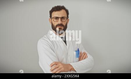 Ein bärtiger Mann mittleren Alters in einem weißen Labormantel mit überkreuzten Armen steht vor einem isolierten weißen Hintergrund. Stockfoto