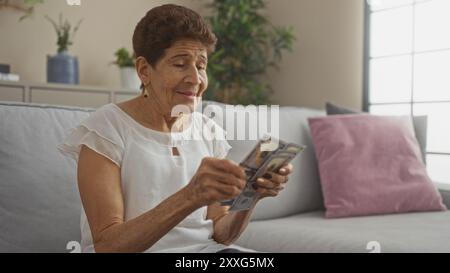 Eine ältere hispanische Frau zählt uns Dollar, während sie auf einer Couch in einem gut beleuchteten Wohnzimmer sitzt und eine ruhige und komfortable Atmosphäre bietet. Stockfoto