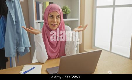 Eine junge Frau mit einem Hijab, die in einem hellen Heimbüro an ihrem Laptop herumschaut. Stockfoto