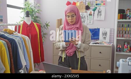 Muslimische Schneiderin mit Tablette in ihrem modernen Atelier, umgeben von bunten Kleidungsstücken und Nähzubehör. Stockfoto