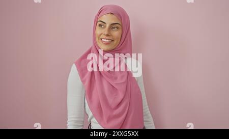 Eine lächelnde junge Frau in einem rosa Hijab vor einem isolierten rosa Hintergrund strahlt Selbstvertrauen und Schönheit aus. Stockfoto