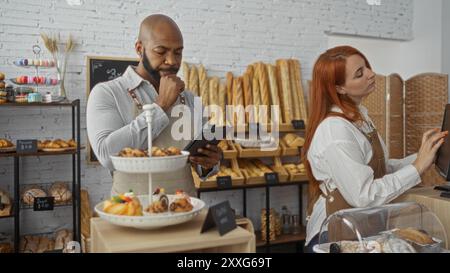 Mann und Frau arbeiten zusammen in einer Bäckerei, umgeben von Gebäck und Brot, tragen Schürzen und verwenden ein Tablet und einen Touchscreen in einem modernen Innenbereich Stockfoto