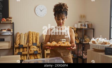 Frau, die Croissants in einer gemütlichen Bäckerei mit Brotregalen und Gebäck hält, eine braune Schürze trägt und drinnen unter einer Uhr steht Stockfoto