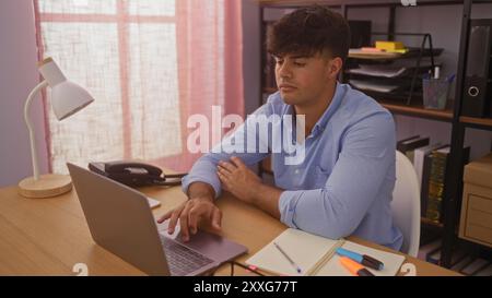 Ein hübscher junger hispanischer Mann in einem Büro, der an einem Laptop arbeitet, während er in einem gut beleuchteten Raum von Bürobedarf und Möbeln umgeben ist. Stockfoto