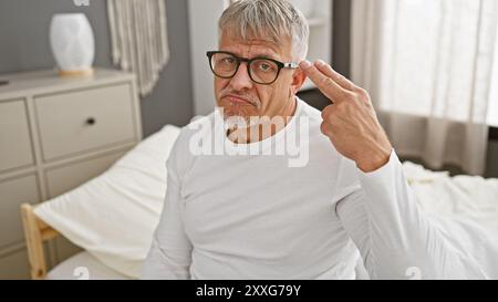 Ein verblüffter, reifer Mann in Brille, der drinnen auf einem Bett sitzt und mit der Hand Unentschlossenheit ausdrückt. Stockfoto