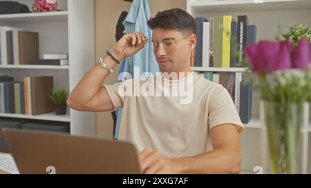 Hübscher hispanischer Mann, der in einem mit Büchern geschmückten Zimmer Brillen anpasst, während er an einem Laptop arbeitete. Stockfoto