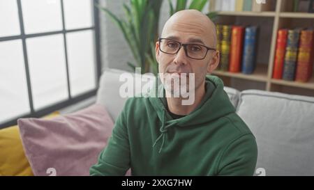 Hübscher Glatzkopf mit Bart, Brille und grünem Hoodie, der drinnen auf der Couch mit Bücherregalhintergrund sitzt Stockfoto