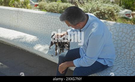 Ein hispanischer Mann mittleren Alters sitzt draußen auf einer gekachelten Bank in einem Stadtpark und streichelt an einem sonnigen Tag zwei kleine Chihuahuas. Stockfoto