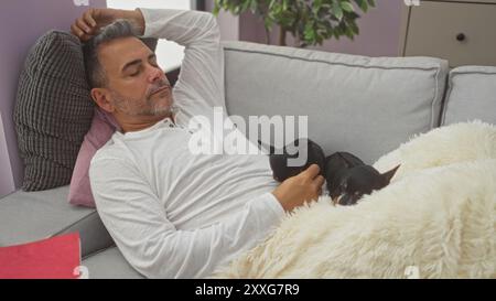 Ein hispanischer Mann mittleren Alters schläft friedlich auf einer Couch in seinem Wohnzimmer und kuschelt zwei kleine Chihuahuas unter einer gemütlichen Decke. Stockfoto