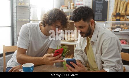 Männer schauen gemeinsam in einer Bäckerei auf Telefone, genießen Kaffee und Gebäck, demonstrieren Kommunikation und Technologie in einer entspannten Umgebung. Stockfoto