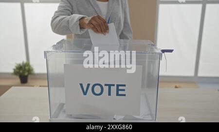 Eine afroamerikanische Frau legt ihren Wahlzettel in eine transparente Wahlbox in einem Innenraum und betont die Bedeutung des Bürgerengagements A Stockfoto