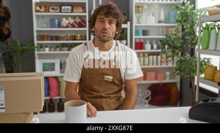 Ein junger Mann mit Bart und brauner Schürze sitzt in einem Haus-Dekorationsgeschäft, umgeben von verschiedenen Innendekorationen und Zimmerpflanzen. Stockfoto