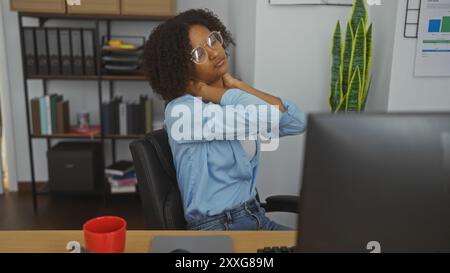 Lockige junge Frau mit Brille, die Nackenschmerzen in einer Büroumgebung mit Computer und roter Tasse auf dem Schreibtisch spürt Stockfoto