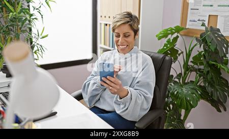 Eine lächelnde hispanische Frau mit kurzen blonden Haaren nutzt ihr Smartphone gern in einer modernen Büroumgebung. Stockfoto