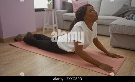 Eine junge afroamerikanische Frau mit lockigen Haaren übt Yoga in einem Wohnzimmer, dehnt sich auf einer rosa Matte, trägt ein weißes Hemd und eine schwarze Hose. Stockfoto