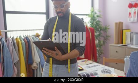 afroamerikanischer Schneider macht Notizen in einem bunten Schneidergeschäft, umgeben von Kleidung und Designmaterialien. Stockfoto