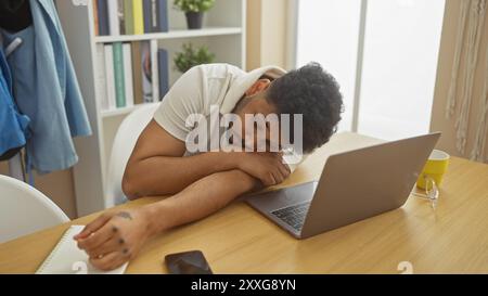 Ein müder afroamerikaner, der an seinem Schreibtisch schläft, mit einem Laptop, Telefon und einer Brille in der Nähe. Stockfoto