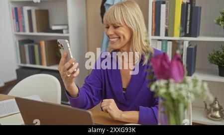 Eine lächelnde blonde Frau in einem lila Pullover genießt es, ihr Smartphone zu Hause zu verwenden und schafft so eine lässige und angenehme Innenumgebung. Stockfoto