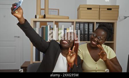 Zwei afroamerikanische Frauen machen ein Selfie, während sie lächeln und in einem Büro sitzen, mit Regalen im Hintergrund Stockfoto