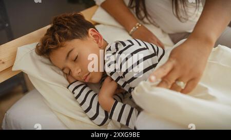 Ein kleiner Junge schläft friedlich auf einem Bett, während ihn eine Frau sanft hineinsteckt und einen zärtlichen Familienmoment im Haus darstellt. Stockfoto