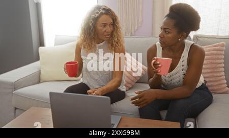 Zwei Frauen, die sich in einem Wohnzimmer entspannen und dabei Tassen halten, während sie sich unterhalten. afroamerikanische und lateinamerikanische Freunde saßen auf einem Sofa in der Nähe Stockfoto