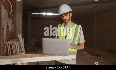 Junger hispanischer Mann auf einer Baustelle, der Sicherheitsausrüstung trägt und einen Laptop mit zielgerichtetem Ausdruck benutzt Stockfoto