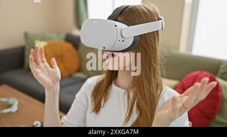 Eine junge blonde Frau erlebt die virtuelle Realität in Innenräumen und drückt ihr Staunen in ihrem Wohnzimmer aus. Stockfoto