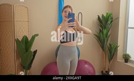 Junge Frau, die Selfie im Fitnessraum mit Fitnessgeräten und Pflanzen macht, einen schwarzen Sport-BH und graue Leggings trägt, blondes Haar, lächelnd und Urlaub Stockfoto