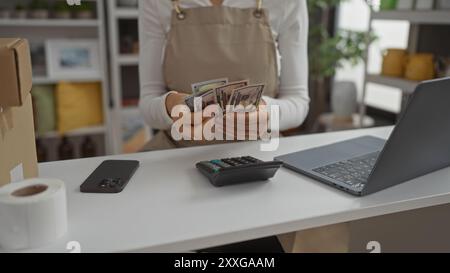 Frau zählt Geld in einem Dekorationsgeschäft, mit Händen, die uns Dollar zeigen, neben einem Laptop und Taschenrechner auf einem Tisch, umgeben von Dekorationsgegenständen Stockfoto