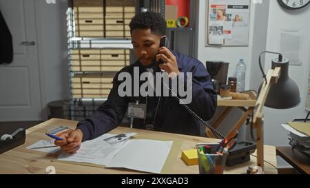 Ein männlicher afroamerikanischer Detective arbeitet an seinem Schreibtisch mit Dokumenten und einem Telefon in einem Polizeirevier. Stockfoto