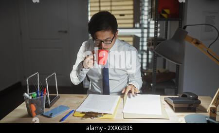 Ein asiatischer Mann schlürft Kaffee, während er spät in einem überfüllten Büro arbeitet, was ein Gefühl von Hingabe vermittelt. Stockfoto